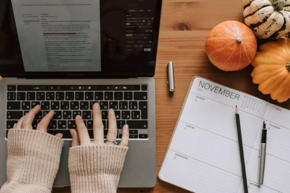 hands typing on a laptop. Beside the laptop is a notebook, pens, and fall decors.